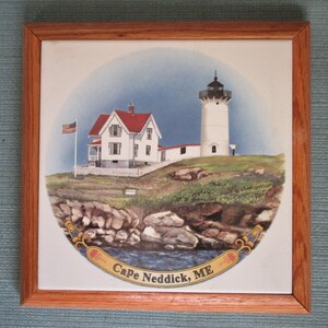 May include: A framed ceramic tile featuring a lighthouse scene. The artwork depicts a white lighthouse with a black top, a white house with a red roof, and an American flag. The text "Cape Neddick, ME" is displayed on the tile.