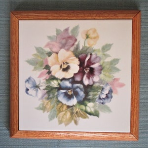 May include: Square decorative tile with a floral design. The tile features a bouquet of pansies in shades of yellow, purple, and blue, surrounded by green leaves. The tile is set in a light brown wooden frame.