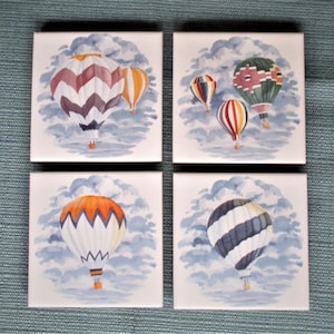 May include: Four square ceramic tiles with watercolor paintings of hot air balloons in a cloudy sky. Each tile features a different color scheme and design of hot air balloons.