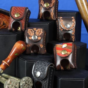 May include: A collection of leather tool holders with various designs, including a US flag, leaves, and a geometric pattern. The holders are displayed on black surfaces with a wooden mallet and a leather carving.