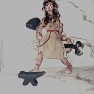Könnte beinhalten: Eine handgefertigte Figur eines Schmieds mit braunem Haar, bekleidet mit einer beigen Tunika, die mit einer dünnen Kordel gebunden ist. Die Figur hält einen dunkelgrauen Hammer in der einen und eine schwarze Metallzange in der anderen Hand.