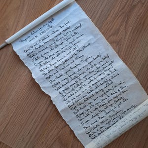 May include: A scroll with handwritten text, held by two silver rods. The text appears to be a prayer or incantation, with phrases like "Come to me, O beloved mistress." The paper has a torn edge, giving it an aged appearance.