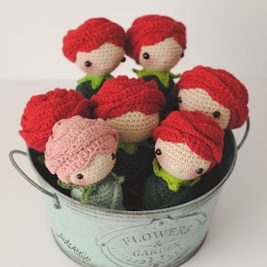 Op de afbeelding: Een verzameling van zes gehaakte rozenpoppen in een blauwe metalen emmer met de tekst "Flowers & Garden" aan de zijkant. De poppen dragen allemaal groene bladeren en hebben rood en roze gehaakt haar.