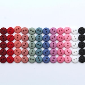 Peut inclure: Une collection de 48 boutons colorés dans différentes nuances de rouge, rose, violet, bleu, vert et noir. Les boutons sont disposés en rangée, les couleurs passant du rouge au noir.