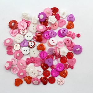 以下が含まれることがあります： ハート、花、リボンなど、さまざまな形をしたカラフルなプラスチックボタンのコレクション。ボタンはピンク、赤、白の色合いです。