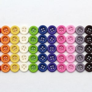 Könnte beinhalten: Eine Regenbogen-Auswahl an Plastikknöpfen in verschiedenen Farben, darunter Rot, Orange, Gelb, Grün, Blau, Lila, Rosa, Grau, Braun und Weiß.
