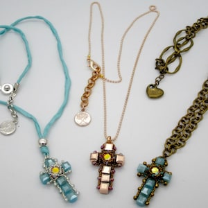 May include: Three necklaces with cross pendants. One has a light blue cord and a silver cross pendant with blue and yellow accents. Another has a gold chain and a cross pendant with pink and yellow accents. The third has a bronze chain and a blue and yellow cross pendant.