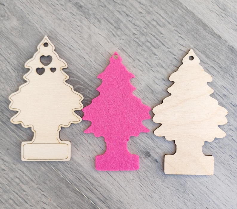 Puede incluir: Tres adornos de &aacute;rbol de Navidad. Dos son de madera clara, uno con recortes de coraz&oacute;n. El tercero es un &aacute;rbol de fieltro rosa vibrante. Todos tienen un agujero en la parte superior para colgar y una base rectangular.