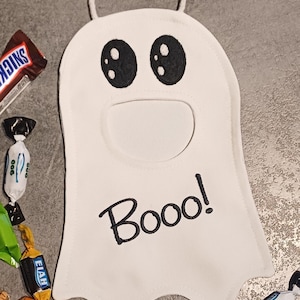 May include: A white ghost-shaped Halloween treat bag with black embroidered eyes and the word "Booo!" in black. The bag has a white handle and is surrounded by wrapped candies.