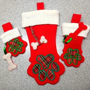 May include: Red Christmas stockings with white faux fur trim and paw print designs. The stockings feature a plaid pattern in the paw prints and bone-shaped embellishments. Green ribbon accents add a festive touch.