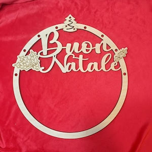 Può includere: Una ghirlanda natalizia in legno con la frase italiana "Buon Natale" intagliata in corsivo. La ghirlanda presenta un piccolo albero di Natale e foglie di agrifoglio. La ghirlanda è circolare e presenta piccoli fori sul bordo.