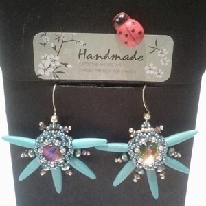 Może przedstawiać: Ręcznie robione kolczyki o kwiatowym wzorze. Kolczyki mają centralną iryzującą koralik otoczoną małymi niebieskimi i srebrnymi koralikami, z jasno niebieskimi akcentami w kształcie płatków. Widoczna jest również mała biedronka i etykieta z napisem "Handmade".