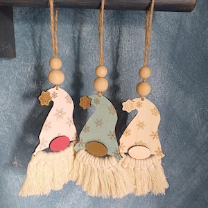 Puede incluir: Tres adornos de gnomos de madera con patrones de copos de nieve y barbas de macramé. Cada gnomo tiene un sombrero de diferente color: rosa, azul y madera natural. Los adornos están suspendidos por hilo y cuentas de madera, ideales para la decoración navideña.