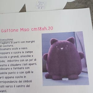 May include: A plush, purple cat toy with a heart-shaped nose and simple facial features. The toy is approximately 7 inches tall and 7 inches wide. The image also shows text in Italian, including the dimensions of the toy.
