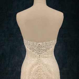May include: A white lace wedding dress with a button-up back. The dress is on a mannequin and the background is a dark blue color.
