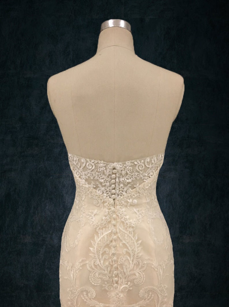 May include: A white lace wedding dress with a button-up back. The dress is on a mannequin and the background is a dark blue color.