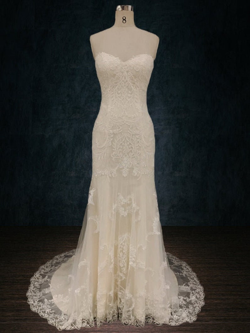 May include: A white strapless wedding dress with lace detailing and a mermaid silhouette. The dress has a sweetheart neckline and a long train.