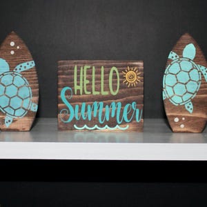 May include: Wooden decorative set featuring two surfboard-shaped pieces with turquoise sea turtle designs and a square block with "HELLO SUMMER" text. The set is made of stained wood.