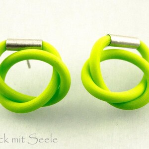Op de afbeelding: Een paar neon limoengroene oorknopjes met een geknoopt lusontwerp, vastgezet door een cilindervormig zilverkleurig metalen accent. De tekst Schmuck mit Seele staat in de linkerbenedenhoek van de afbeelding.