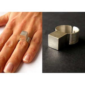 May include: A silver-toned ring featuring a prominent, solid cube-shaped centerpiece attached to a wide, flat band. The metal surface displays a brushed, matte texture throughout the entire structure, highlighting the geometric design of the piece.