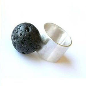 Könnte beinhalten: Ein Ring mit einer breiten, glatten, polierten Silberschiene, die an einem einzelnen, kugelförmigen, porösen schwarzen Vulkangestein befestigt ist. Der Kontrast zwischen dem metallischen Glanz des Silbers und der dunklen, strukturierten Oberfläche des Steins prägt das Schmuckstück.