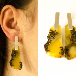 Könnte beinhalten: Ein Paar Bernstein-Ohrringe mit silbernen Stiften. Der Bernstein ist gelb und hat eine raue, natürliche Textur. Die Ohrringe sind ein einzigartiges und stilvolles Schmuckstück.