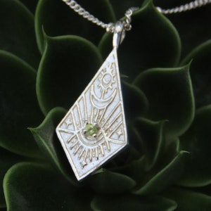 Może przedstawiać: Srebrny naszyjnik z geometrycznym wisiorem, w którym znajduje się zielony kamień szlachetny w środku. Wisiorek ma kształt diamentu i jest ozdobiony wygrawerowanym słońcem, księżycem i gwiazdami.