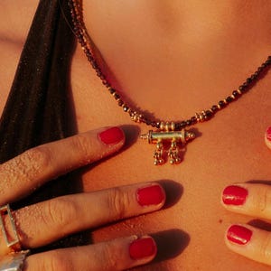 May include: A gold pendant necklace with a beaded chain. The pendant has a cylindrical design with dangling elements. The image also shows red nail polish and silver rings.