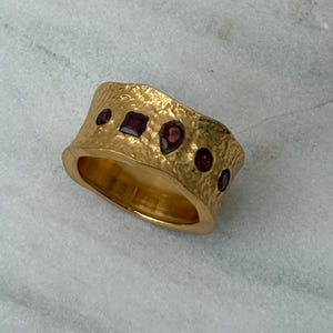 May include: A gold ring with a textured surface and a wide band. The ring is set with small, dark red gemstones. The ring has a hammered finish. The gemstones are of different shapes. The ring has a handcrafted appearance.