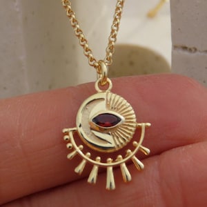 Puede incluir: Un collar dorado con un colgante que presenta una luna creciente, un diseño de rayos de sol y una pequeña gema roja ovalada. El colgante cuelga de una delicada cadena. El collar se muestra sobre un fondo neutro.