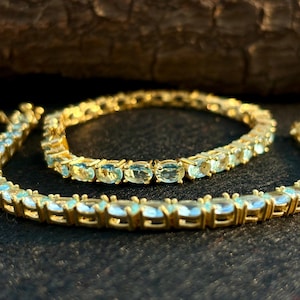 Op de afbeelding: Goudkleurige armbanden met ovale, lichtblauwe edelstenen. De armbanden worden gepresenteerd op een donkere ondergrond, met een vage houten achtergrond. De edelstenen zijn dicht op elkaar gezet, waardoor een doorlopende lijn van kleur ontstaat.