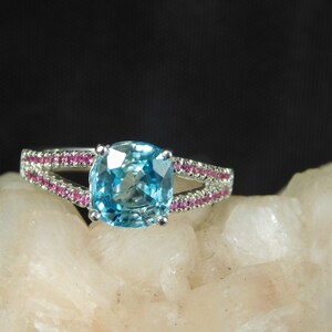 May include: A silver ring with a large, cushion-cut blue topaz gemstone surrounded by a double row of pink sapphires.