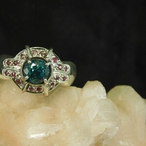 May include: A silver ring with a large, round, teal blue gemstone surrounded by a halo of smaller, pink gemstones.