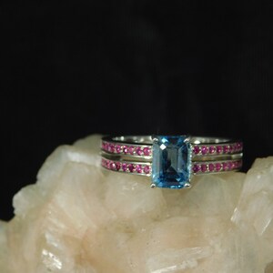 May include: A silver ring with a rectangular, light blue gemstone as the centerpiece. Two bands of smaller, pink gemstones flank the main stone. The ring is set against a neutral, textured background.