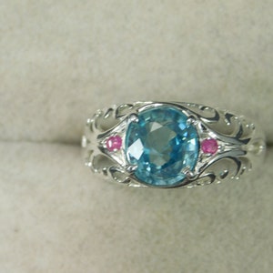 May include: A silver ring with a blue oval gemstone and two smaller pink gemstones. The ring has a delicate, ornate design.
