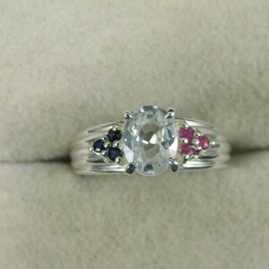 May include: A silver ring with a large clear oval gemstone surrounded by smaller blue and red gemstones.
