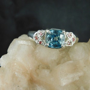 May include: A silver ring with a blue gemstone surrounded by smaller pink gemstones.