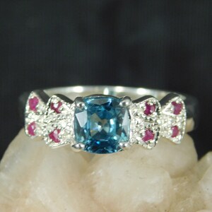 May include: A silver ring with a blue gemstone surrounded by smaller red gemstones. The ring has a delicate design with a bow-like shape on each side of the center stone.