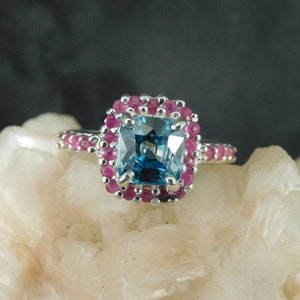 May include: A silver ring featuring a large, square, blue gemstone surrounded by smaller, round, pink gemstones. The ring's band is also adorned with pink gemstones. The ring is set on a white, textured surface.