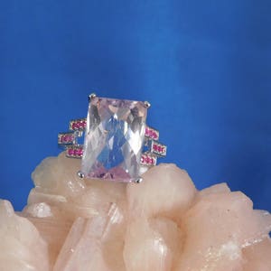 May include: A pink gemstone ring with a rectangular cut stone surrounded by a silver band with small pink gemstones.