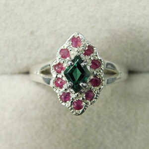 Puede incluir: Anillo de plata con una piedra central verde oscuro tallada en esmeralda, rodeada de pequeñas piedras rojas redondas. El anillo tiene un diseño ovalado alargado y una banda dividida.