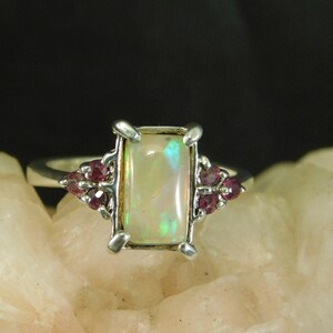 Puede incluir: Un anillo de plata con una piedra de ópalo rectangular y pequeñas piedras rosadas en la banda.