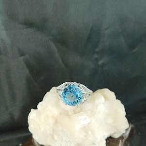 Może przedstawiać: Srebrny pierścionek z dużym, okrągłym, niebieskim topazem otoczonym mniejszymi, przezroczystymi kamieniami.