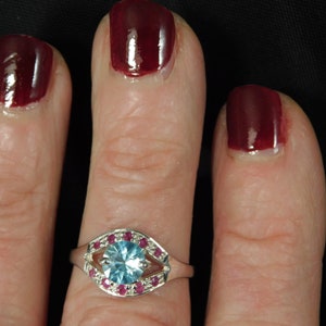 May include: A silver ring with a large light blue gemstone surrounded by a halo of smaller pink gemstones.