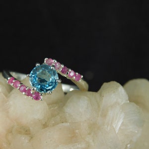 May include: A silver ring with a blue topaz gemstone surrounded by pink sapphires.