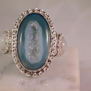 Puede incluir: Un anillo de plata con una gran piedra ovalada de ágata azul con un centro blanco, flanqueada por dos piedras blancas redondas más pequeñas. El anillo tiene un borde plateado trenzado alrededor de la piedra principal.