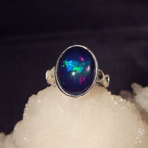 May include: A silver ring with an oval, dark blue opal gemstone. The opal has iridescent flashes of green, blue, and pink. The ring is set on a white, textured surface against a dark background. The ring's band has a decorative design.