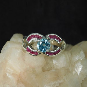 May include: A silver ring with a blue gemstone surrounded by small red gemstones.