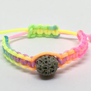 May include: A multi-colored braided bracelet with a dark gray lava rock bead. The bracelet is made with neon pink, yellow, green, and blue cords.
