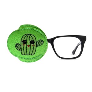 May include: A green felt eye patch with an embroidered cactus design. The cactus has a smiling face with black eyes and a red mouth. The eye patch is attached to a pair of black framed glasses.